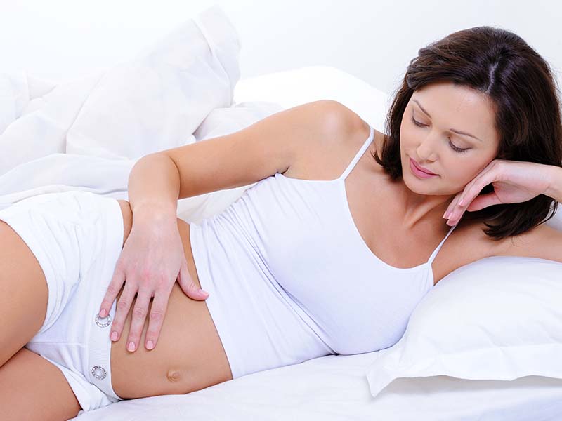 Mulher grávida deitada de lado em uma cama branca, olhando e passando a mão em sua barriga