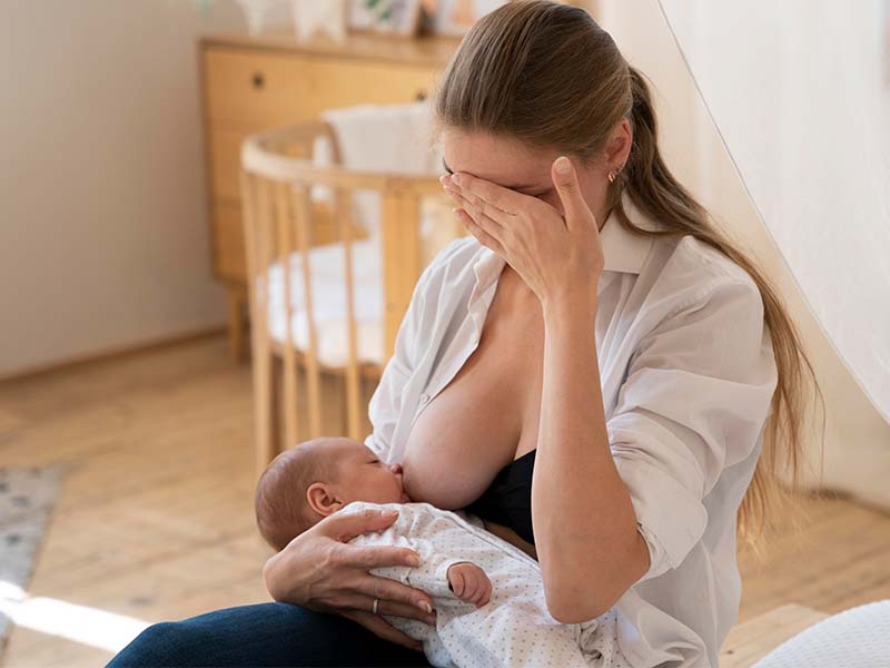 Mãe amamentando seu filho em um quarto e segurando sua própria cabeça, demonstrando dor nos seios durante a amamentação