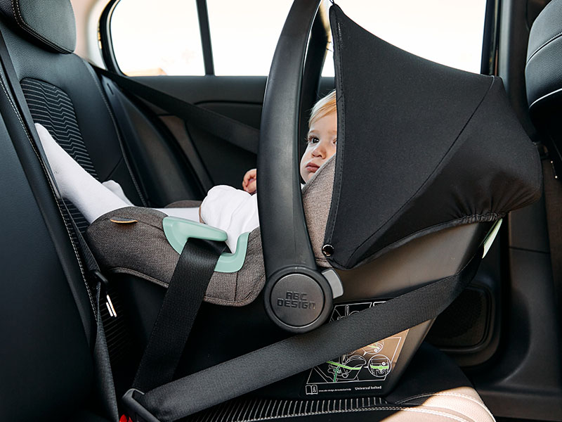 A neném loirinha está de roupa branca, deitada dentro do bebê conforto Tulip Herb da ABC Design de cor marrom e capota preta. O bebê conforto Tulip está preso pelo cinto de segurança do carro.