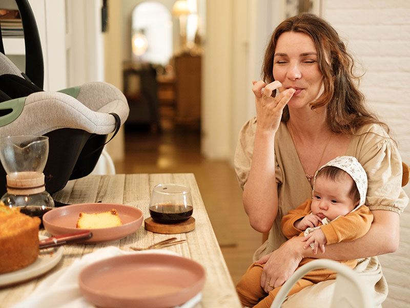 Uma mãe com roupa bege e seu filho no colo, com touca branca e roupinha laranja, sentada próxima a uma mesa com bolo e café, além de um Bebê Conforto Tulip da ABC Design Brasil.
