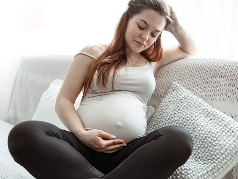 Mulher grávida com roupas claras, sentada em um sofá com tecido e almofadas claras. Uma de suas mãos apoiando a cabeça e a outra por baixo da barriga, sentido as movimentações do seu bebê.