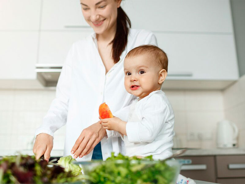 Bebê sentado sem cima da mesa junto com sua mãe, que está preparando a comida, com alimentos saudáveis.