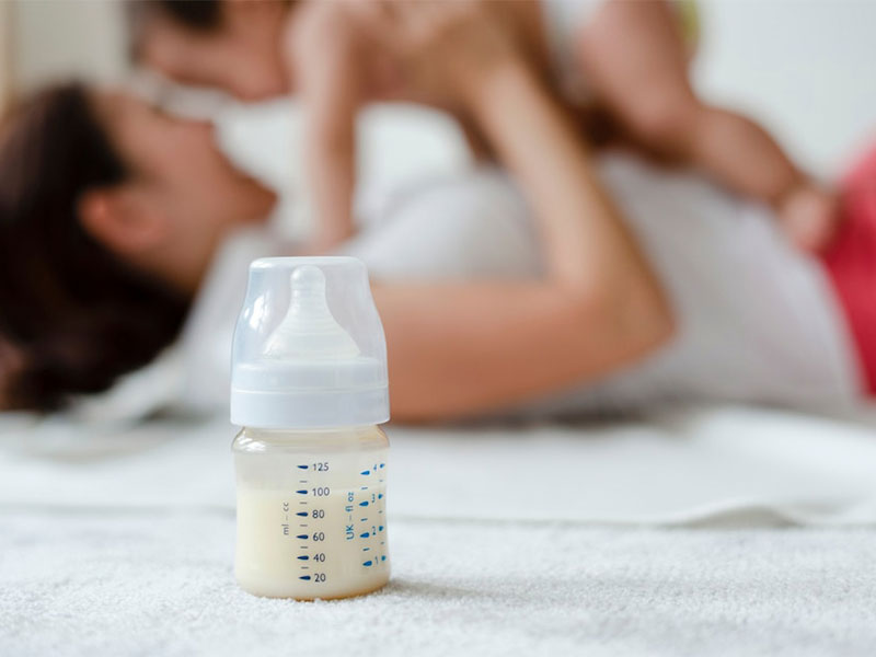 Uma mamadeira com leite em primeiro plano e uma mãe brincando com seu bebê ao fundo desfocado