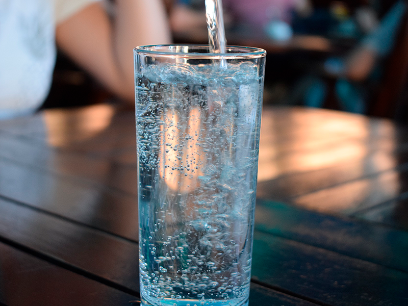 Um copo de vidro com água sendo enchido em cima de uma mesa de madeira em um lugar movimentado.