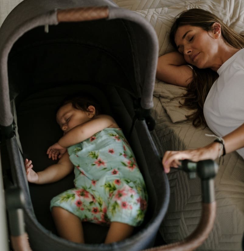Mãe dormindo na cama, e bebê ao lado no carrinho dormindo
